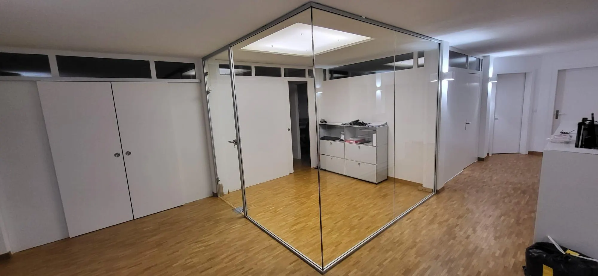 Rahmenlose Ganzglas-Trennwand über Eck auf Parkett, im Hintergrund ein weisser Schrankraum